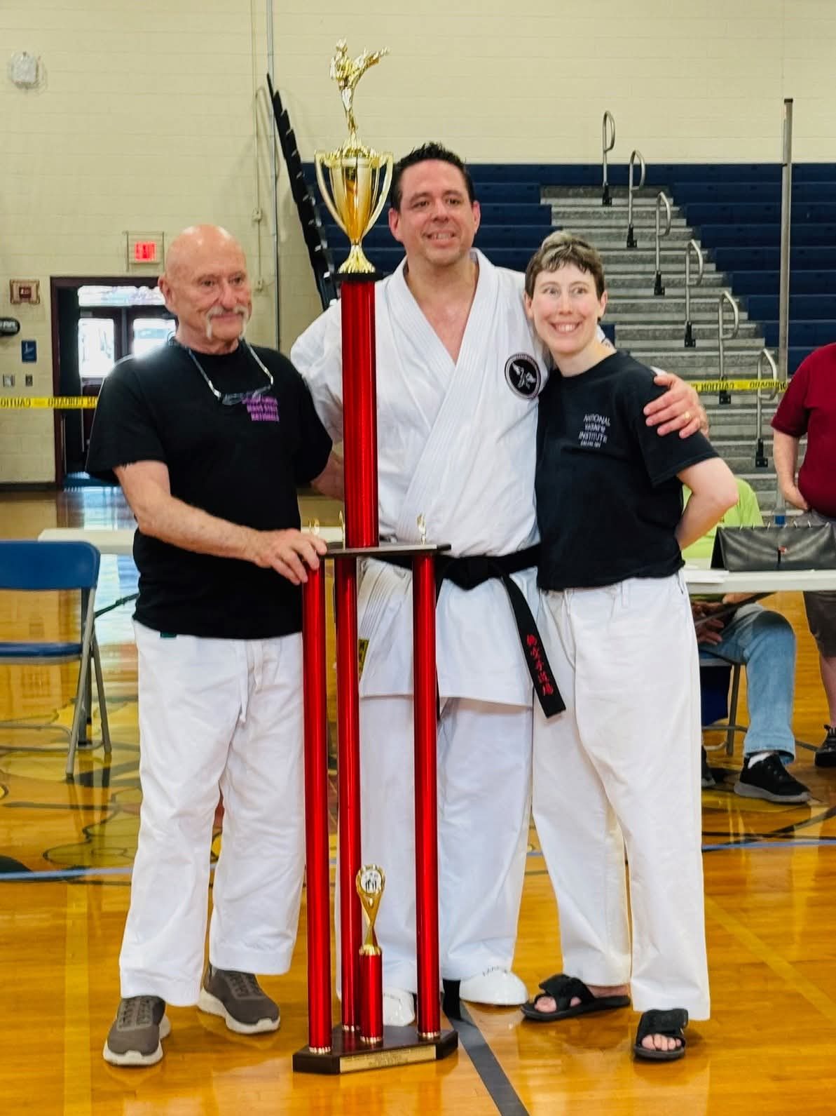 Matt Hogan in his karate gi and black belt, holding a grand champion trophy alongside two fellow martial artists